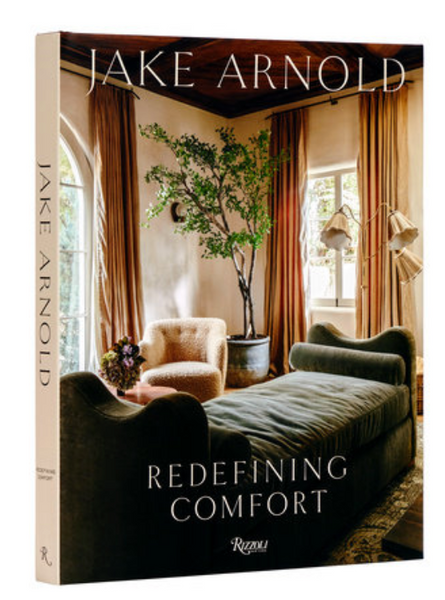 The cover of "Jake Arnold: Redefining Comfort" by Rizzoli features a cozy, well-furnished living room scene epitomizing warm minimalism, with elegant seating, a green chaise lounge, and large windows draped with curtains.