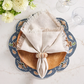 A wooden plate rests on a blue floral placemat with a beige fringed napkin held by the Kim Seybert Shell Arch Napkin Ring, alongside two empty glasses and a vase of white flowers—ideal for coastal entertaining.