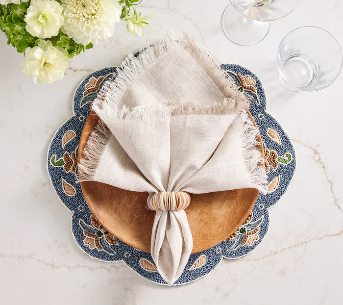 A wooden plate rests on a blue floral placemat with a beige fringed napkin held by the Kim Seybert Shell Arch Napkin Ring, alongside two empty glasses and a vase of white flowers—ideal for coastal entertaining.