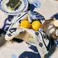 A table set with Johanna Ortiz’s Ikat Amazónico Napkins, a candle, a plate with tinned fish, and a basket holding lemons wrapped in blue and white linen radiates exotic elegance.