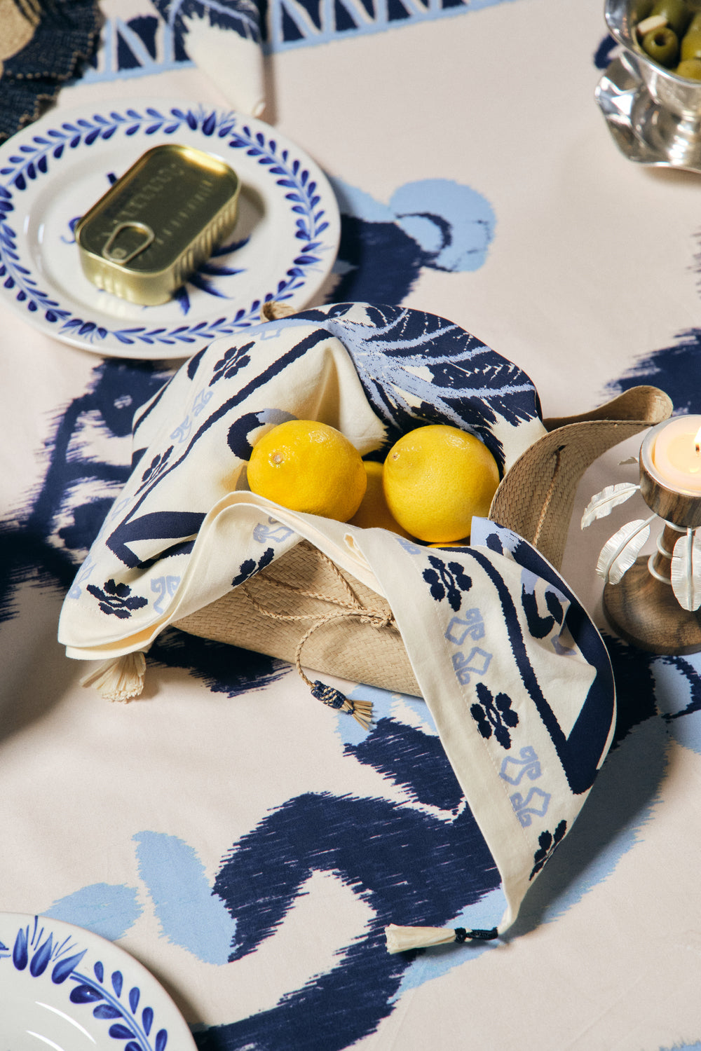 A table set with Johanna Ortiz’s Ikat Amazónico Napkins, a candle, a plate with tinned fish, and a basket holding lemons wrapped in blue and white linen radiates exotic elegance.