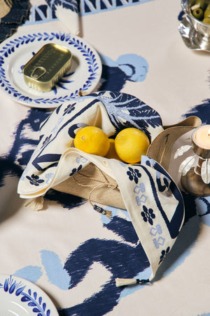 A table set with Johanna Ortiz’s Ikat Amazónico Napkins, a candle, a plate with tinned fish, and a basket holding lemons wrapped in blue and white linen radiates exotic elegance.