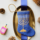 The Bauble Stockings Light the Menorah Stocking by Bauble Stockings features a blue menorah design on a gold platter, elegantly adorned with a blue ribbon. This heirloom-quality stocking is beautifully surrounded by pinecones and colorful dreidels on a white background, setting an ideal scene for honoring family traditions.