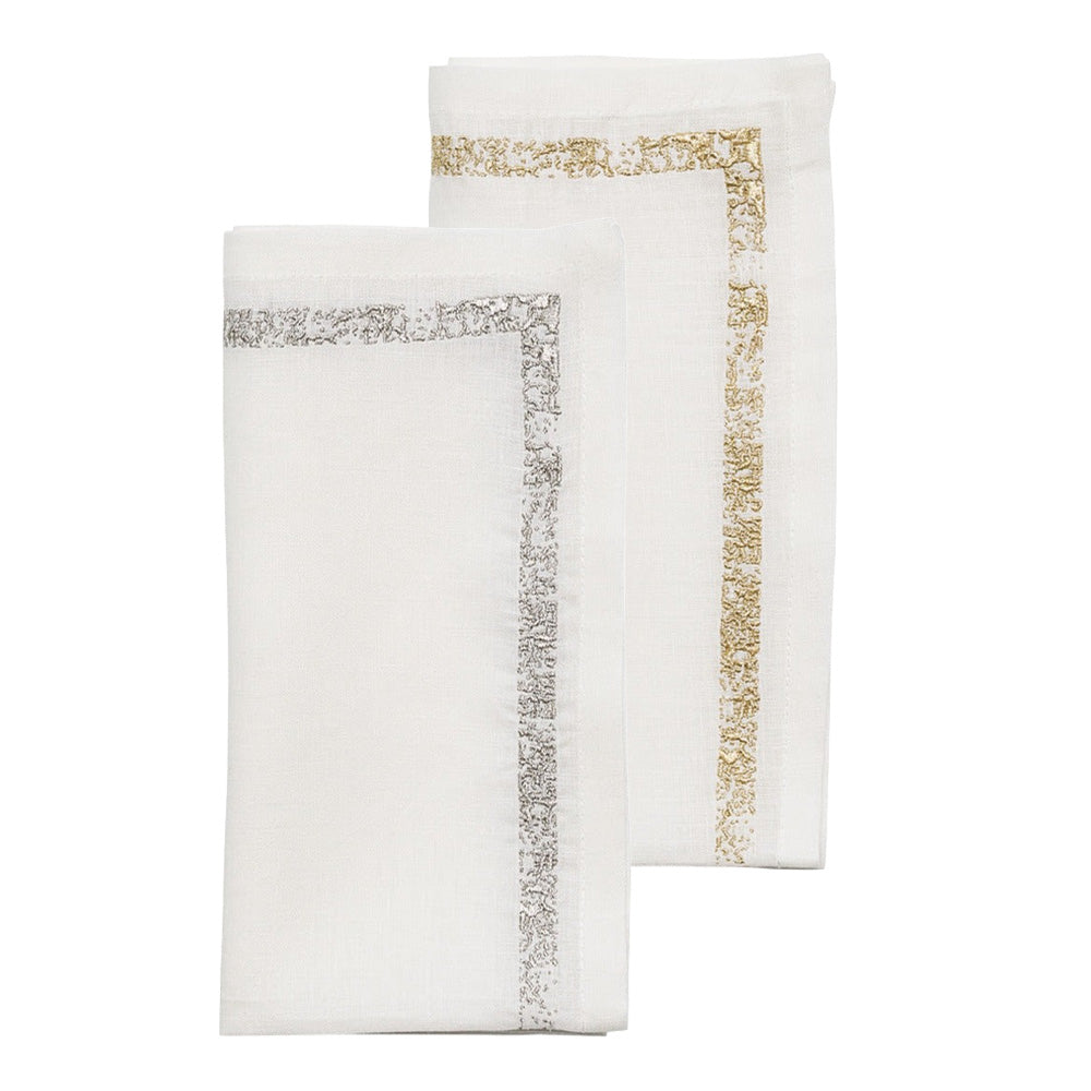 The Kim Seybert Impression Napkin Set includes two neatly folded white linen napkins—one with a silver embroidered border and one with a gold decorative border.