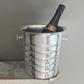 A bottle of sparkling wine is chilled in a Hôtel Silver Vintage Champagne Bucket with Flared Top & Ribbed Body, set on the corner of a distressed wooden surface against a plain wall.