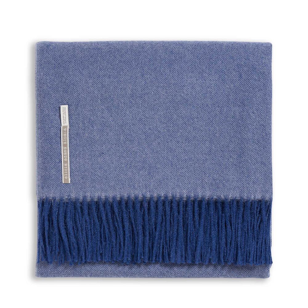 The Classic Herringbone Throw Collection by Alicia Adams Alpaca features a folded blue herringbone-patterned baby alpaca blanket with dark blue fringe, a left-side label, and is fair-trade made in Peru.