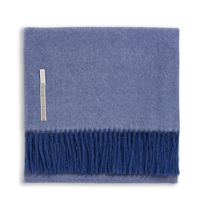 The Classic Herringbone Throw Collection by Alicia Adams Alpaca features a folded blue herringbone-patterned baby alpaca blanket with dark blue fringe, a left-side label, and is fair-trade made in Peru.