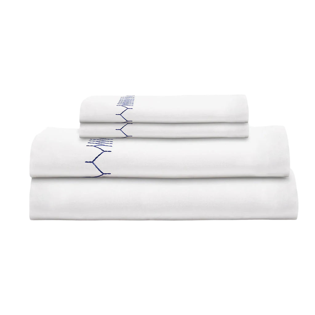A neatly stacked set of John Robshaw’s Stitched Indigo Bedding Collection—white organic cotton sheets with blue stitched detailing—displayed against a plain white background.