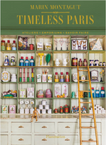 A shelving unit inspired by artisan studios is adorned with colorful jars, boxes, figurines, and artfully placed objects—all arranged as if curated from creative adventures—and features the book “Timeless Paris” by Penguin Random House displayed alongside a wooden ladder for a touch of inspiration.