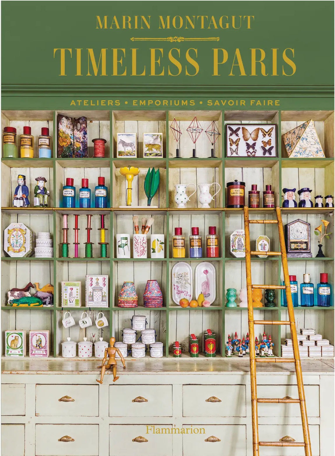 A shelving unit inspired by artisan studios is adorned with colorful jars, boxes, figurines, and artfully placed objects—all arranged as if curated from creative adventures—and features the book “Timeless Paris” by Penguin Random House displayed alongside a wooden ladder for a touch of inspiration.