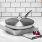 The Hestan Commercial Clad Stainless Steel Universal Lid, 11", rests on a marble surface beside two red onions and a striped cloth, with a white tiled wall as the ideal backdrop for this space-saving kitchen essential.