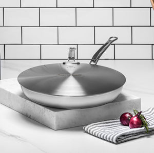The Hestan Commercial Clad Stainless Steel Universal Lid, 11", sits atop a marble slab beside a striped towel and two red onions, all set against a white tiled backdrop.