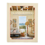The cover of Island Hopping by Common Ground showcases a coastal living room with wicker furniture, beach hats, and an ocean view through an open door—perfectly capturing interiors inspired by the relaxed elegance of island living.