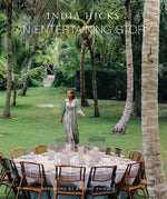 A woman in a floral dress stands on grass beside a round outdoor dining table set with candles and wooden chairs, surrounded by lush green trees—offering perfect party inspiration reminiscent of the relaxed elegance featured in An Entertaining Story by Common Ground.