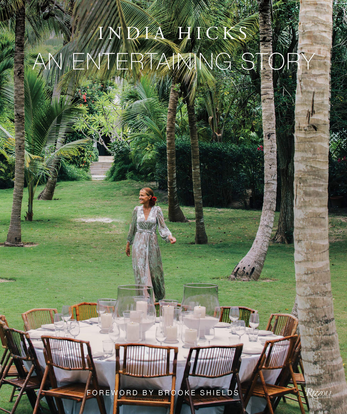 A woman in a floral dress stands on grass beside a round outdoor dining table set with candles and wooden chairs, surrounded by lush green trees—offering perfect party inspiration reminiscent of the relaxed elegance featured in An Entertaining Story by Common Ground.