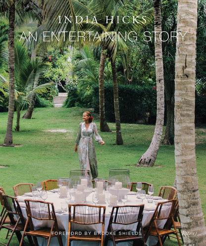 A woman in a floral dress stands on grass beside a round outdoor dining table set with candles and wooden chairs, surrounded by lush green trees—offering perfect party inspiration reminiscent of the relaxed elegance featured in An Entertaining Story by Common Ground.