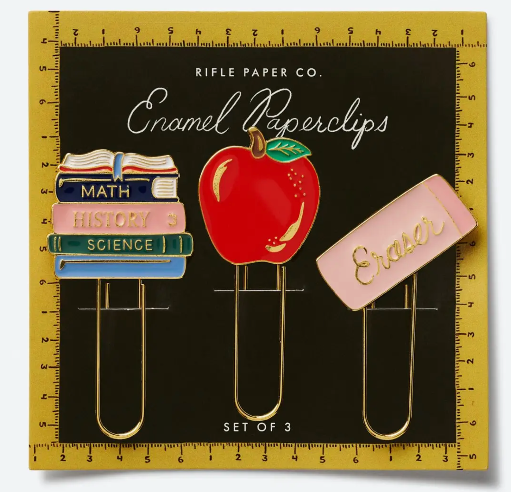 Three enamel paperclips from the Rifle Paper Co. Schoolhouse Collection—shaped like a stack of books, a red apple, and a pink eraser—are displayed on a black card with a gold ruler border, making them ideal for adding charm to your desk supplies or giving as thoughtful teacher gifts.