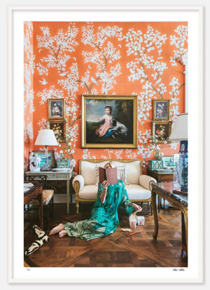 A person in a green dress sits on the floor reading a book in a room with vibrant orange, floral-patterned wallpaper, decorated with classic paintings, a white sofa, and various antique furnishings. Among the artwork is Nick Mele Fine Art Chinoiserie, meticulously printed on archival Canson Baryta fibre rag paper by Nick Mele Photography.