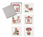 Set of six square white Santa Cocktail Napkins featuring embroidered Christmas-themed scenes, displayed with their gray storage box as elegant holiday party decor.