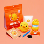 The Woobles Kiki the Chick Beginner Crochet Kit includes yarn, stuffing, a crochet hook, felt pieces, eyes, and step-by-step instructions to make an adorable chick project, all shown on an orange background.