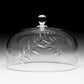 The Jasmine Cake Dome by William Yeoward Crystal features a hand-engraved leaf pattern and a round handle on top, showcased against a gradient backdrop—a true example of handmade glassware.