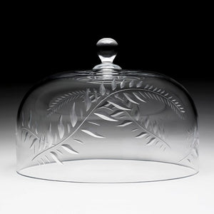 The Jasmine Cake Dome by William Yeoward Crystal features a hand-engraved leaf pattern and a round handle on top, showcased against a gradient backdrop—a true example of handmade glassware.