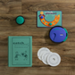 A vintage edition of the Catch Phrase board game titled "Catch Phrase Vintage Bookshelf Edition" by WS Game Company lies on a wooden surface, featuring a book-like box. Inside, you'll find a circular electronic device, a small circular game piece, a rectangular game card, and three game discs.