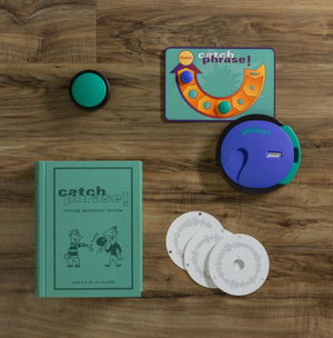 A vintage edition of the Catch Phrase board game titled "Catch Phrase Vintage Bookshelf Edition" by WS Game Company lies on a wooden surface, featuring a book-like box. Inside, you'll find a circular electronic device, a small circular game piece, a rectangular game card, and three game discs.