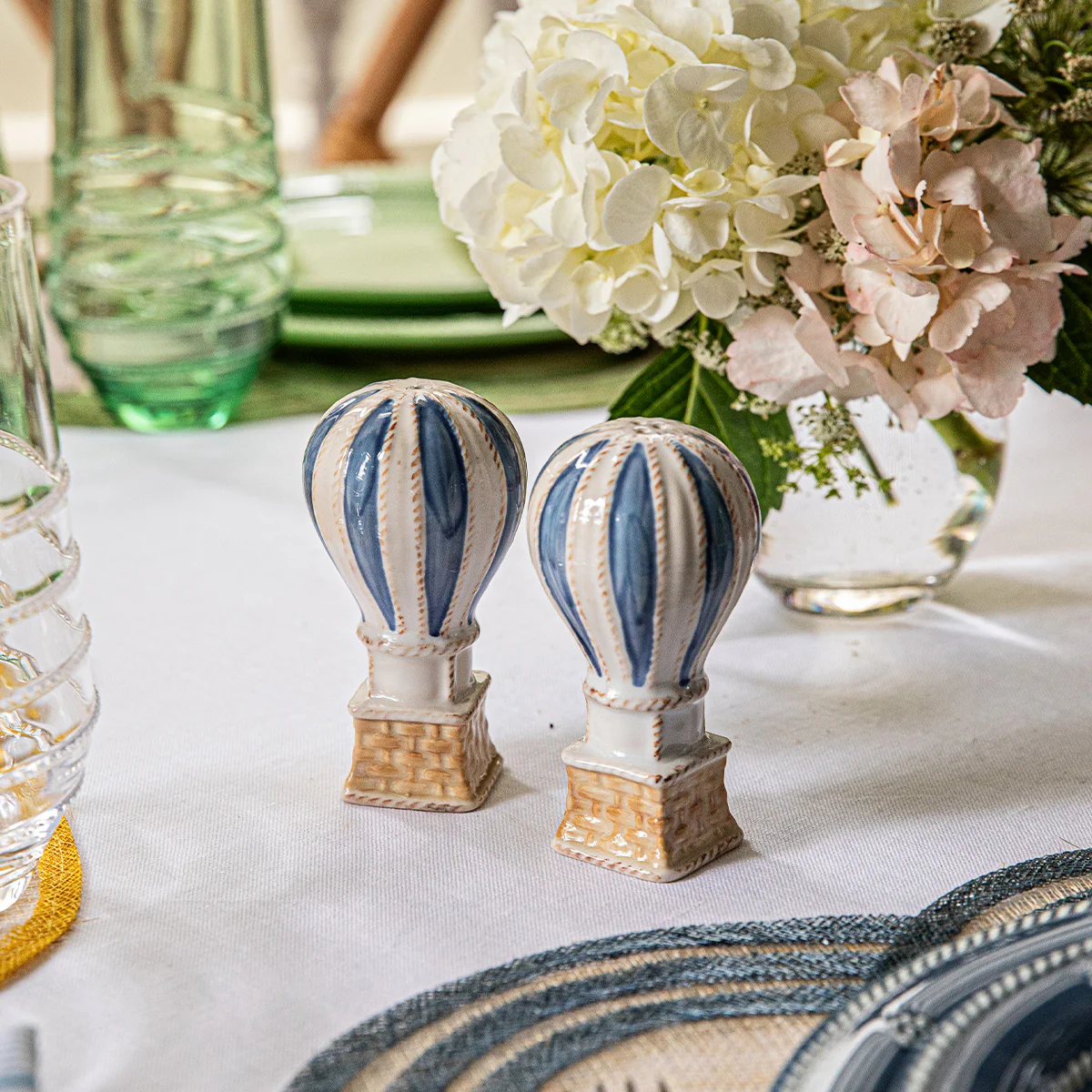 The Juliska L'Amour Toujours Hot Air Balloon Salt and Pepper Shaker Set in blue sits on a dining table with a floral centerpiece and glassware—an enchanting gift, beautifully presented in a Juliska gift box.