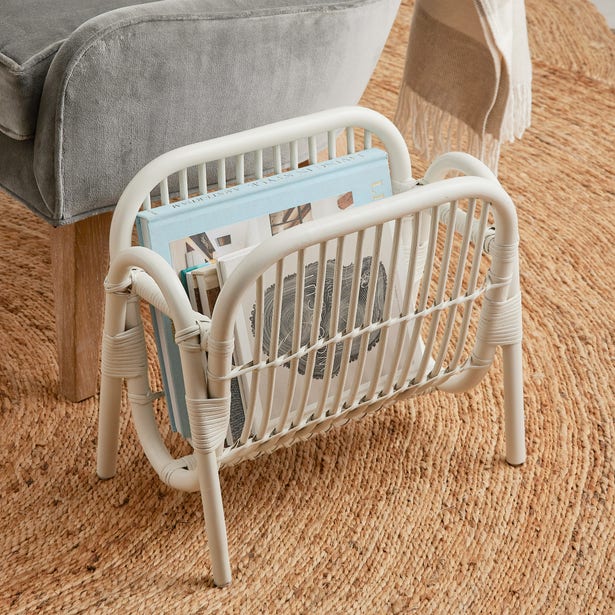 The Makassar Magazine Holder by Pigeon & Poodle, crafted from white rattan, sits on a woven rug beside a gray upholstered chair with a beige blanket, enhancing the charm of your natural decor.