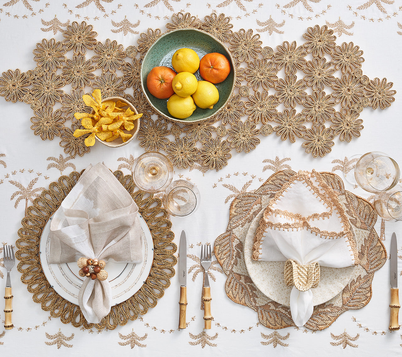 A stylish tablescape with two place settings, gold-trimmed napkins, cutlery, crystal glasses, citrus fruit bowl, and snacks on the Kim Seybert Embroidered Palm Tablecloth adds chic desert flair.