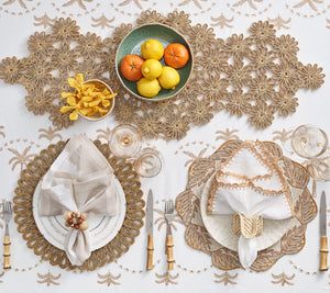 Elegant dining table setting featuring golden placemats, white plates, golden cutlery, and a central bowl of citrus fruits on a Kim Seybert Embroidered Palm tablecloth.