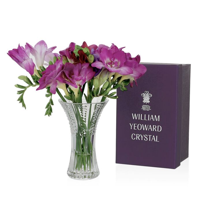 The Julia Posy Vase by William Yeoward Crystal, filled with purple and pink flowers, sits beside a purple gift box—a charming hostess gift.