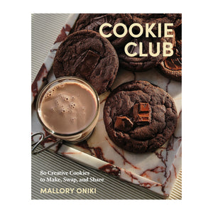 A marble tray holds chocolate cookies and a glass mug of hot chocolate. The book “Cookie Club” by Penguin Random House is featured, hinting at irresistible cookie recipes from this popular cookbook.