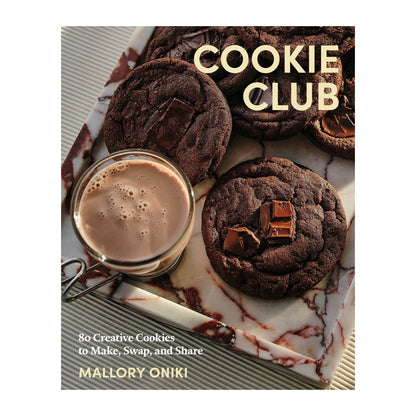 A marble tray holds chocolate cookies and a glass mug of hot chocolate. The book “Cookie Club” by Penguin Random House is featured, hinting at irresistible cookie recipes from this popular cookbook.