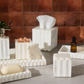 The Proviso White Bath Collection by Pigeon & Poodle features pleated marble tissue boxes, soap dishes, trays, and containers—ideal for luxury decor. Shown on gray with a brown tiled background.