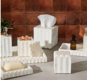 The Proviso White Bath Collection by Pigeon & Poodle features pleated marble tissue boxes, soap dishes, trays, and containers—ideal for luxury decor. Shown on gray with a brown tiled background.