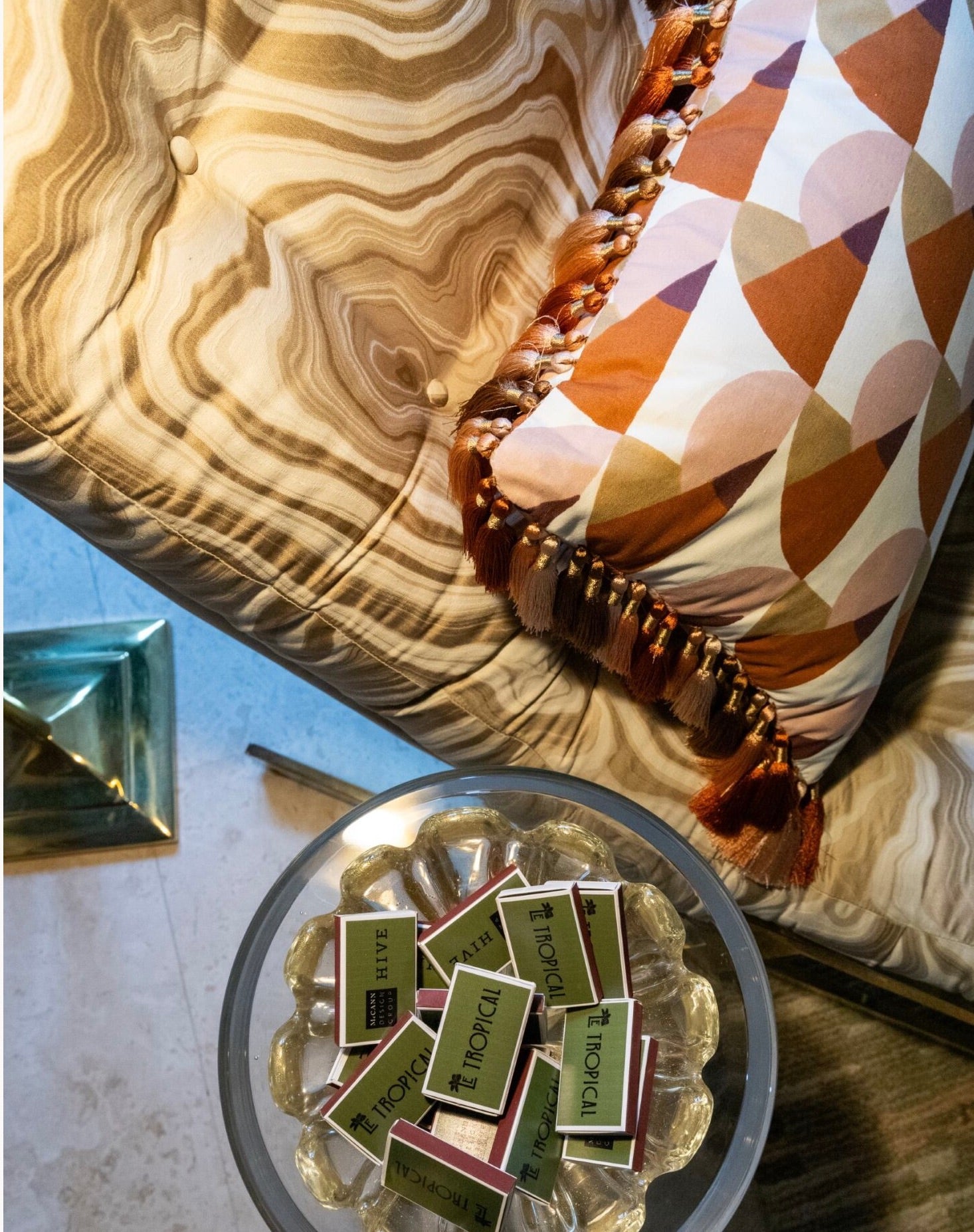 A Manille Cuba Lumbar Pillow with Tassel Fringe by Hive Custom Pillows adds a geometric touch to a tufted sofa beside a round table topped with glass ashtrays and "Tropical" matchboxes, in chic Kips Bay Show House style.