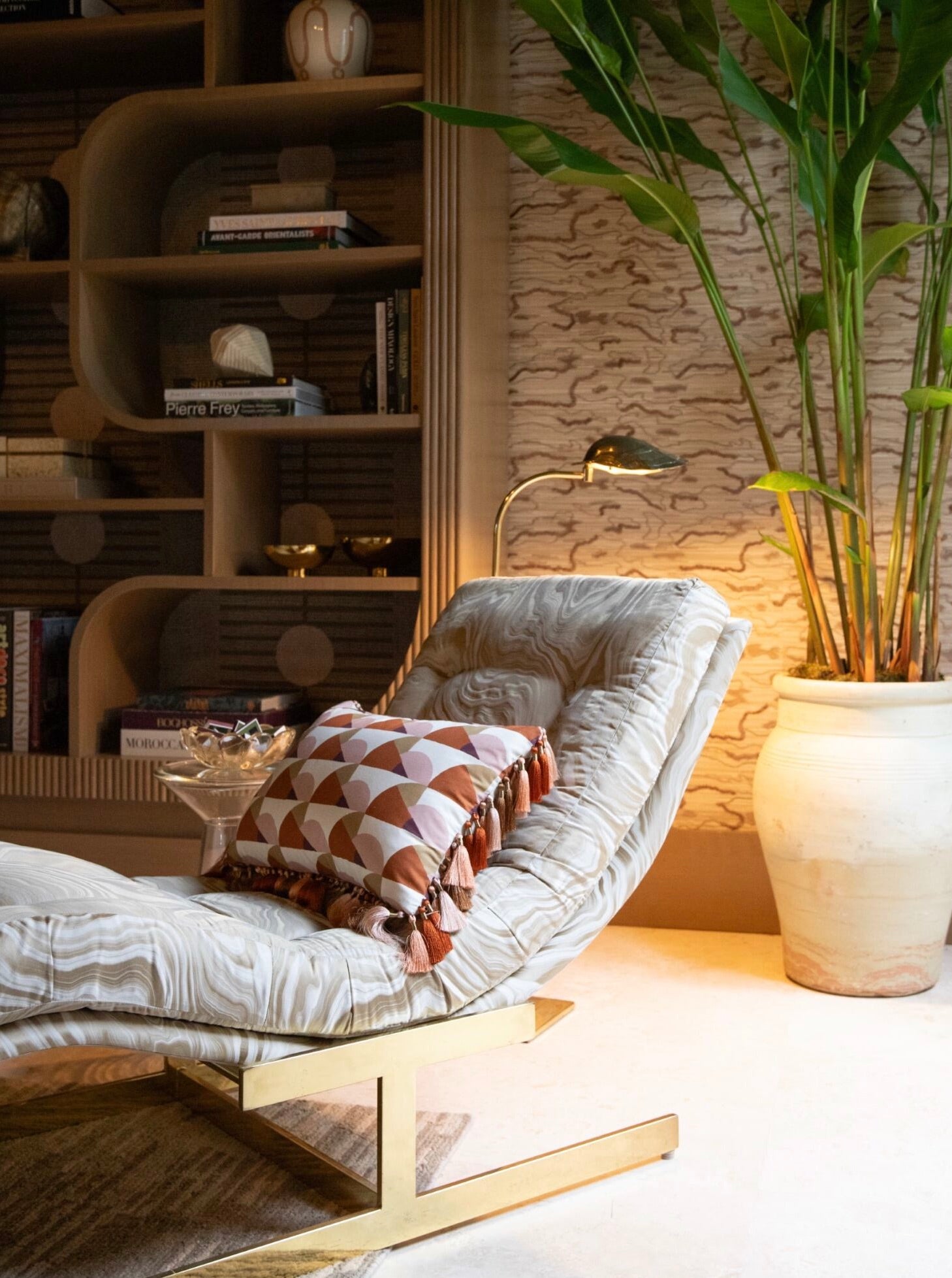 A modern lounge chair with a Manille Cuba Lumbar Pillow with Tassel Fringe by Hive Custom Pillows sits beside a large potted plant and a built-in bookshelf, evoking the chic look of the Kips Bay Show House.