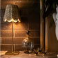 The Poldina Pro Lamp Collection by Zafferano America, featuring cordless illumination and a woven shade, shines next to a glass decanter and glasses on marble, against a dark tile wall.