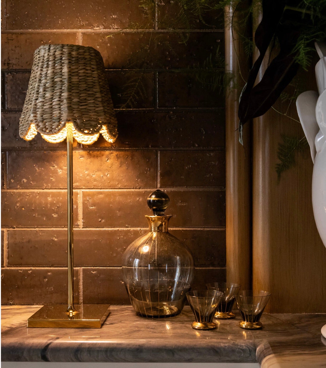 The Poldina Pro Lamp Collection by Zafferano America, featuring cordless illumination and a woven shade, shines next to a glass decanter and glasses on marble, against a dark tile wall.