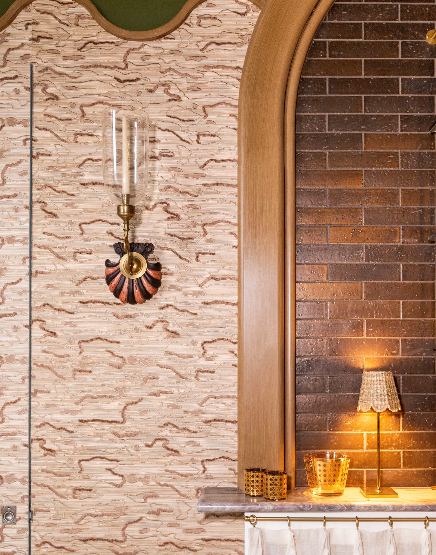 A wall-mounted sconce with a glass shade is paired with the Poldina Pro Lamp from Zafferano America, placed on a marble countertop alongside a glass and basket, in front of dark brown brick tiles and patterned wallpaper.