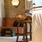 A stylish room reminiscent of the Kips Bay Show House, featuring a bar area with two wooden stools and an ornate bar counter. Behind the bar, there's a brown banquette and unique wall decor, including sculptural pieces and patterned elements. Additionally, the space is accentuated by the inclusion of a Martini Small Ottoman from Associated Design.