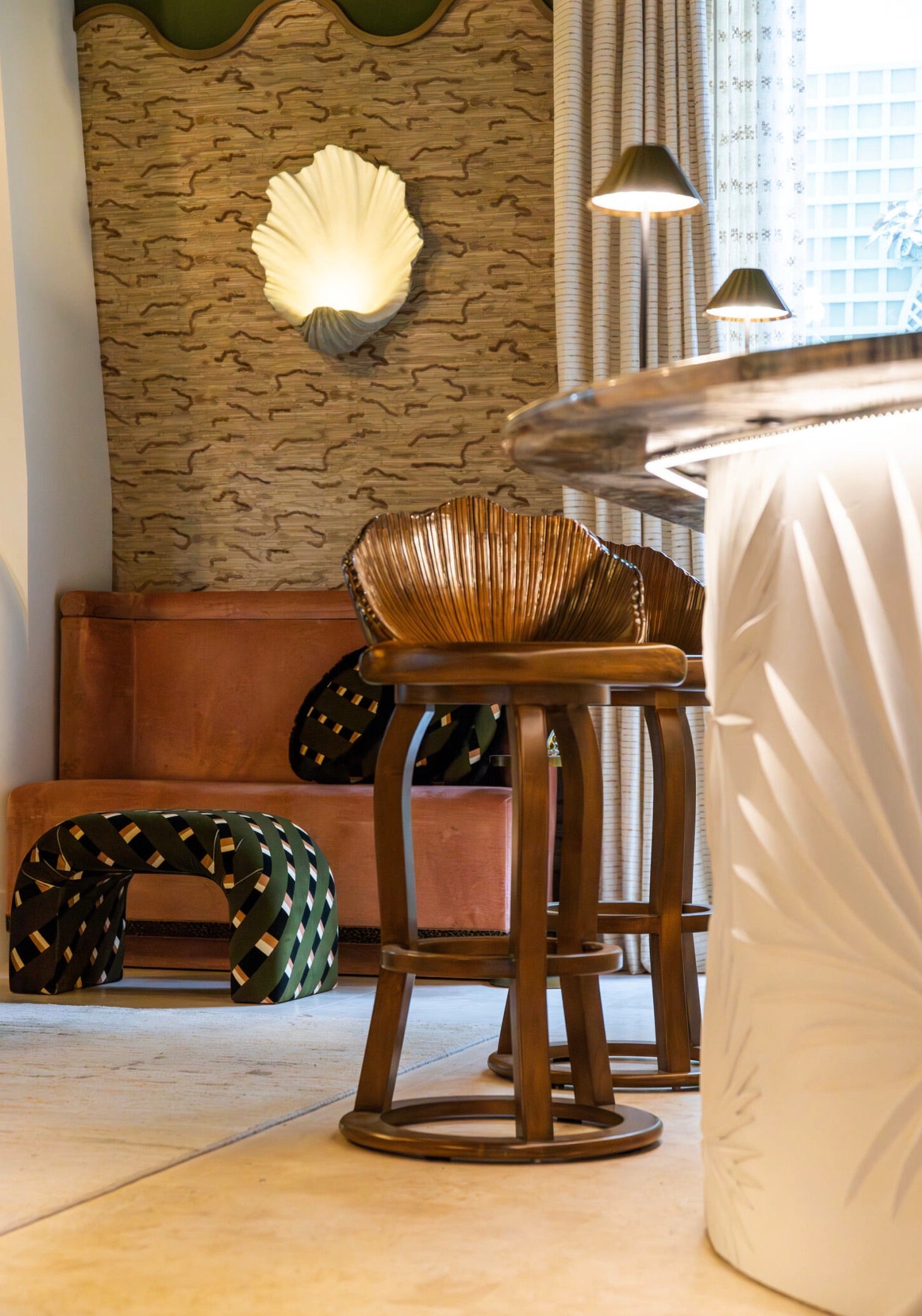 A stylish room reminiscent of the Kips Bay Show House, featuring a bar area with two wooden stools and an ornate bar counter. Behind the bar, there's a brown banquette and unique wall decor, including sculptural pieces and patterned elements. Additionally, the space is accentuated by the inclusion of a Martini Small Ottoman from Associated Design.