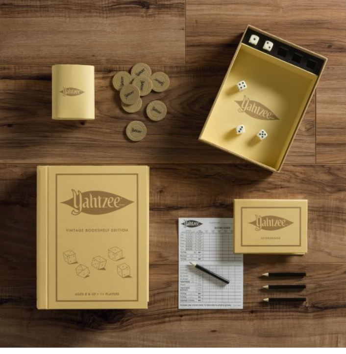 A Yahtzee Vintage Bookshelf Edition set by WS Game Company, featuring dice, scorecards, wooden tokens, and a shaker adorned with vintage-inspired graphics, all displayed on a wooden surface. The box and items are designed in a charming vintage bookshelf edition style.