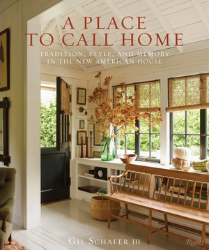 Featuring a bright, welcoming entryway with a wooden bench, potted plant, framed artwork, and an open black door revealing a lush green garden, the cover of A Place To Call Home by Rizzoli showcases the timeless charm that award-winning architect Gil Schafer III brings to inviting home design.