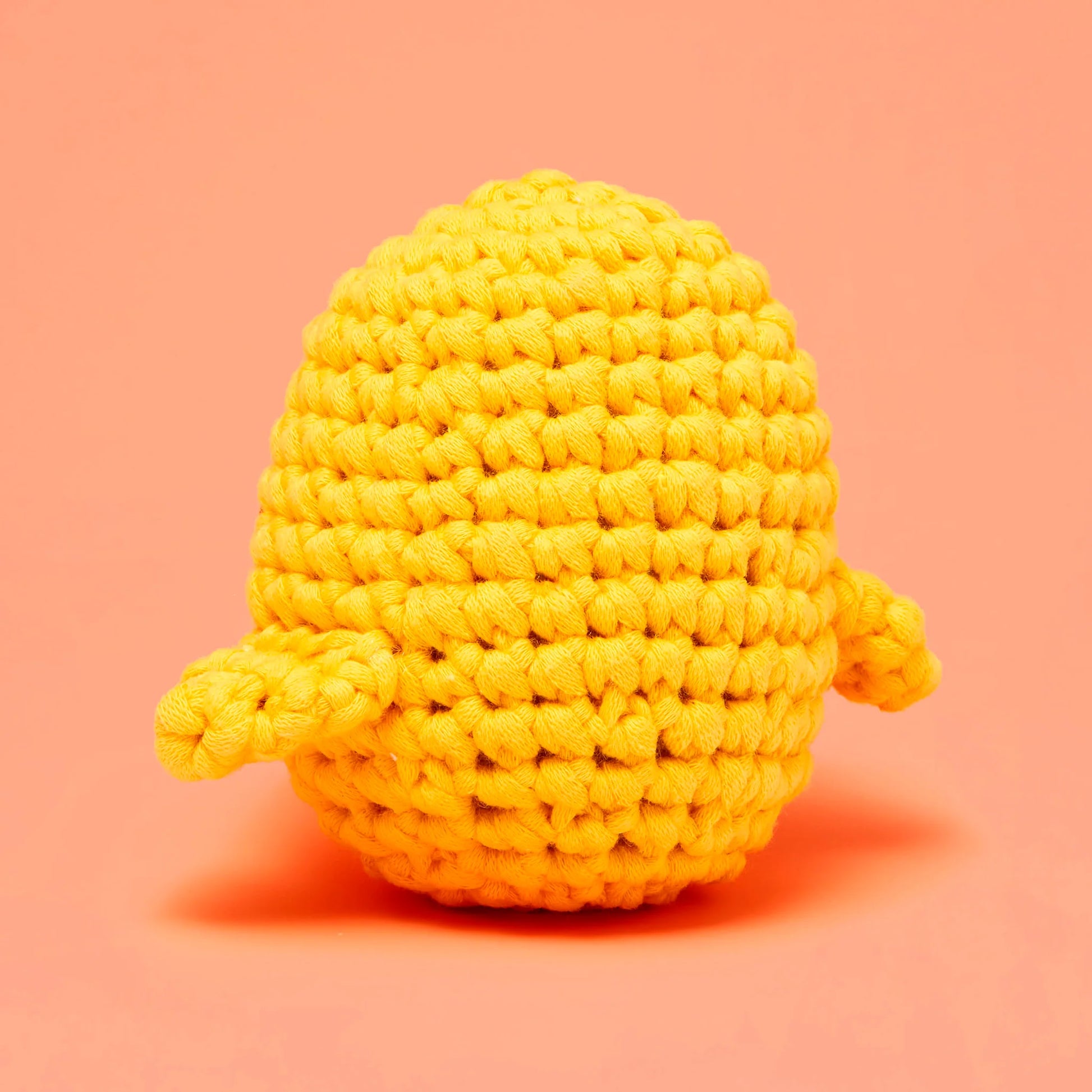 Kiki the Chick Beginner Crochet Kit by The Woobles features a small, round, yellow crochet chick with two short arms, set against a plain peach-colored background—an ideal project for beginner crochet enthusiasts or as part of a complete crochet kit.