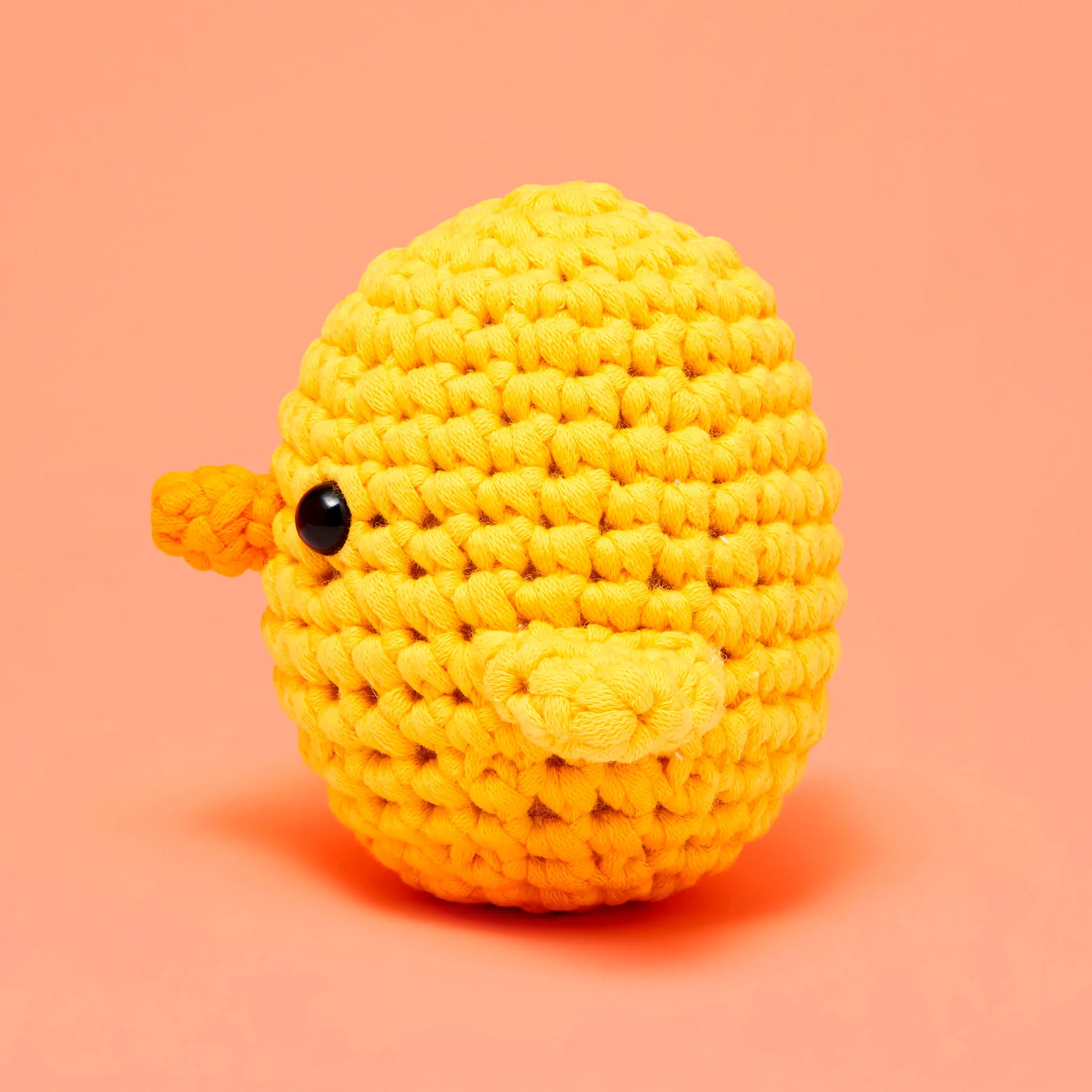 Kiki the Chick Beginner Crochet Kit by The Woobles is a round, yellow, crocheted stuffed toy—ideal for beginner crochet projects—featuring a small black eye and two side flippers, shown against a solid peach background.