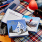 Two open ski books rest on a plaid blanket beside gloves, goggles, a helmet, and skis. One book shows a skier on snowy slopes; the other is "100 Slopes of A Lifetime" by Graphic Image—a must-have ski bucket list.