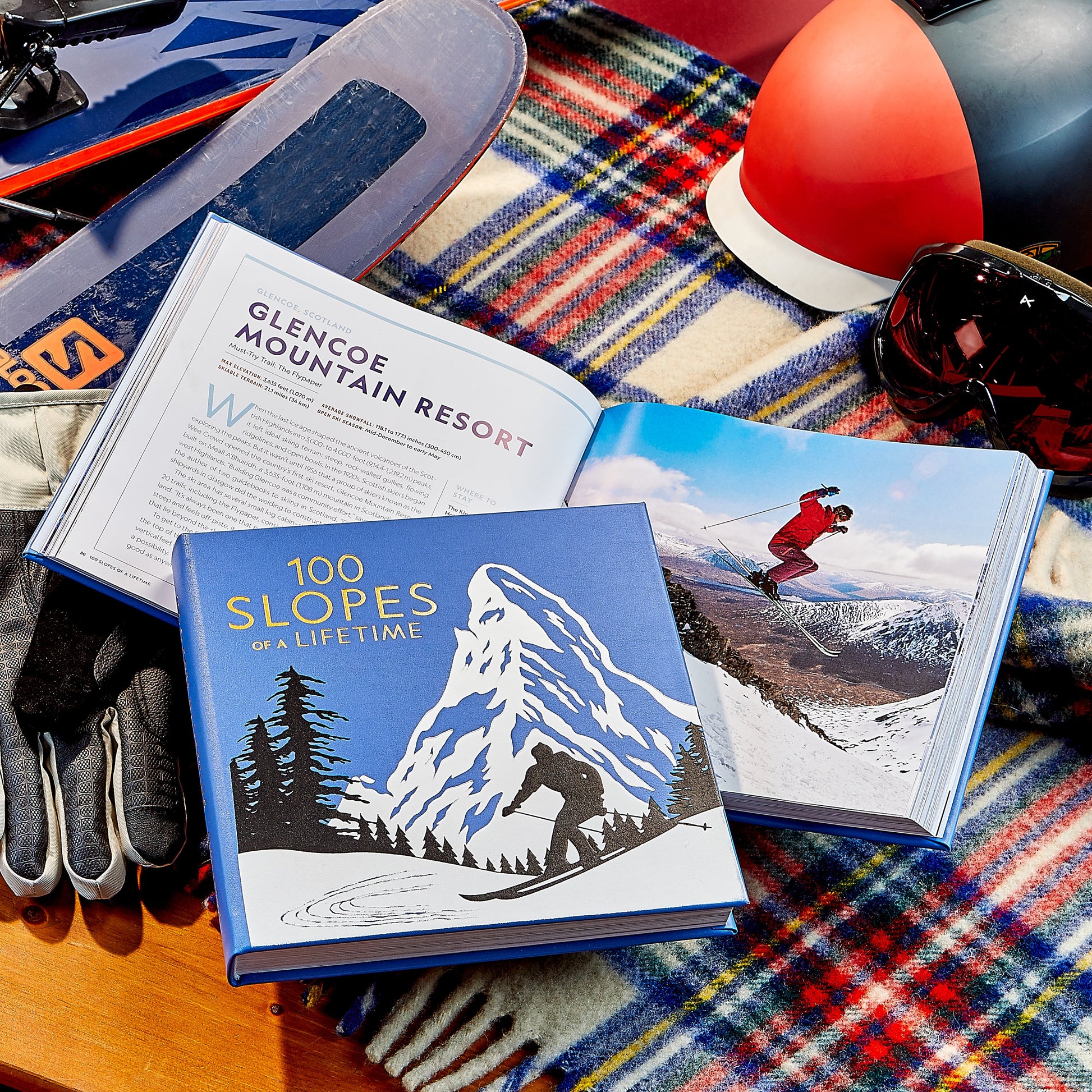 Two open ski books rest on a plaid blanket beside gloves, goggles, a helmet, and skis. One book shows a skier on snowy slopes; the other is "100 Slopes of A Lifetime" by Graphic Image—a must-have ski bucket list.